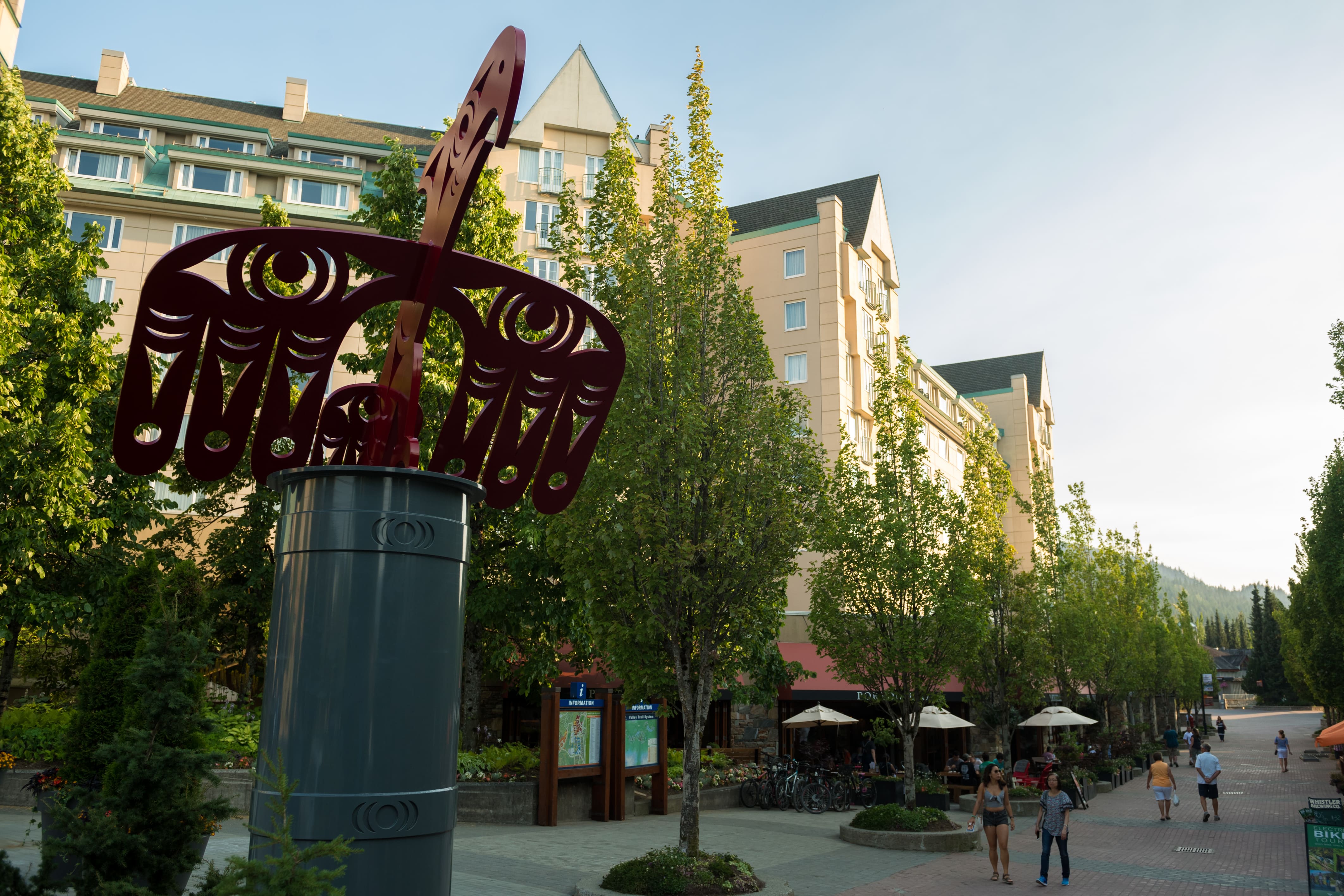 Sculpture in Upper Village with hotel buildings, trees, and people walking in Whistler