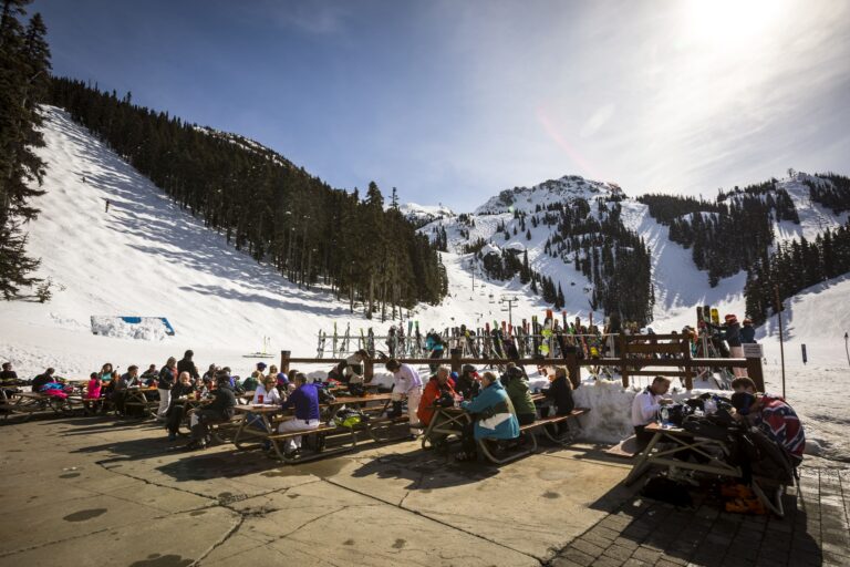 Skiers enjoying food and drinks at an outdoor patio at the base of Whistler Mountain on a sunny winter day