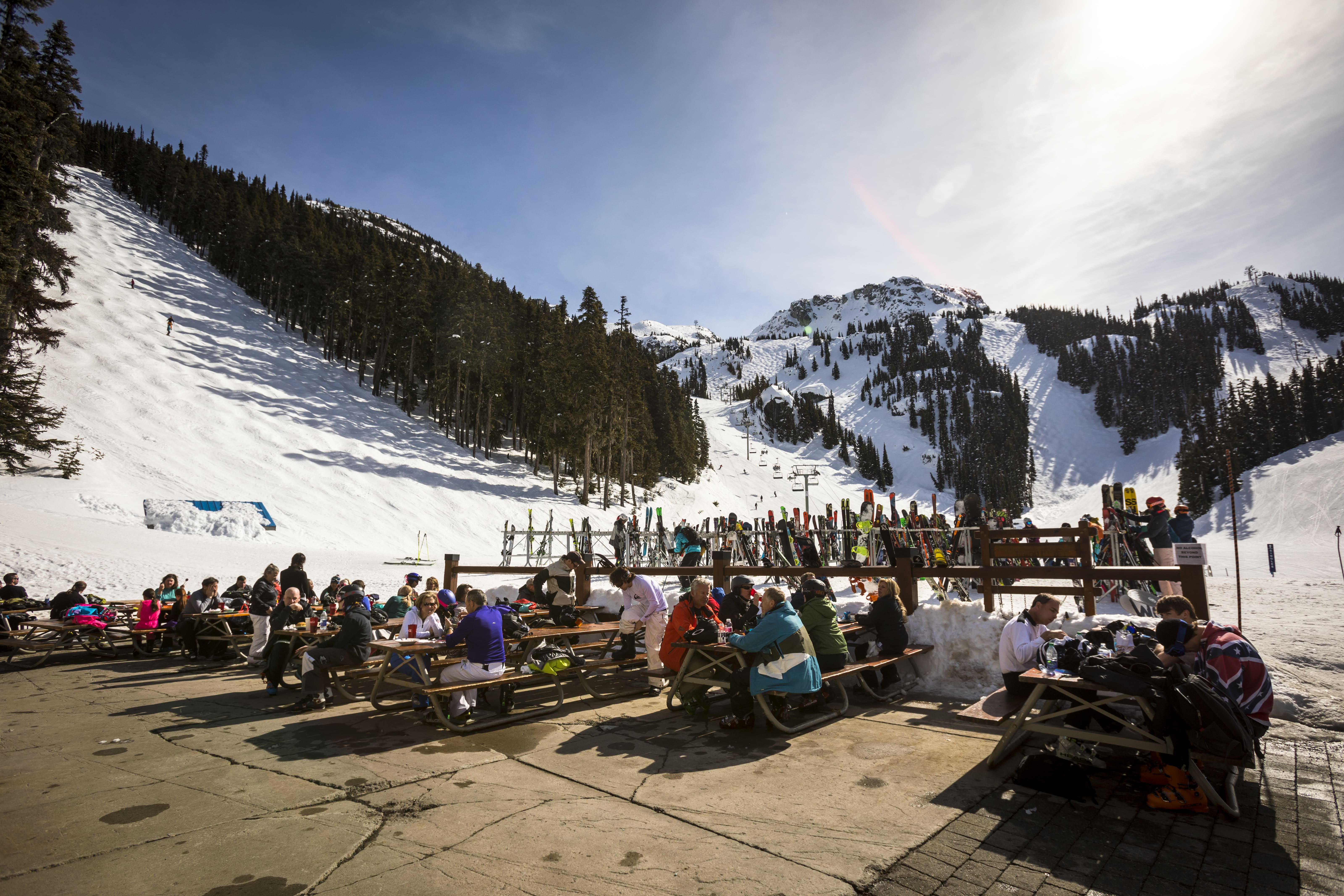 Skiers enjoying food and drinks at an outdoor patio at the base of Whistler Mountain on a sunny winter day